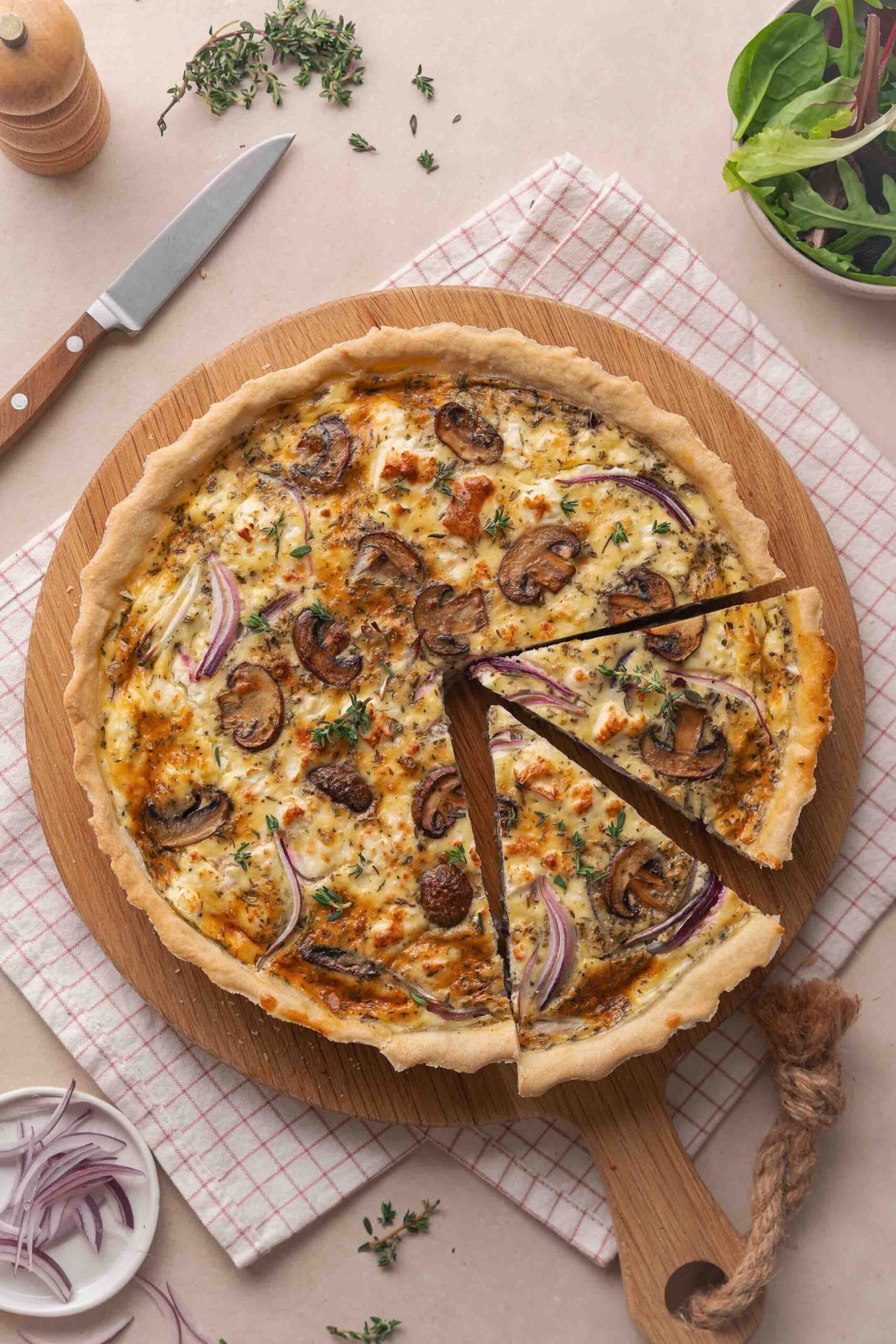 A quiche baked with mushrooms, onions and herbs on a wooden board, with a slice cut out. Nearby are a knife, greens, pepper mill and minced onions on a tea towel.