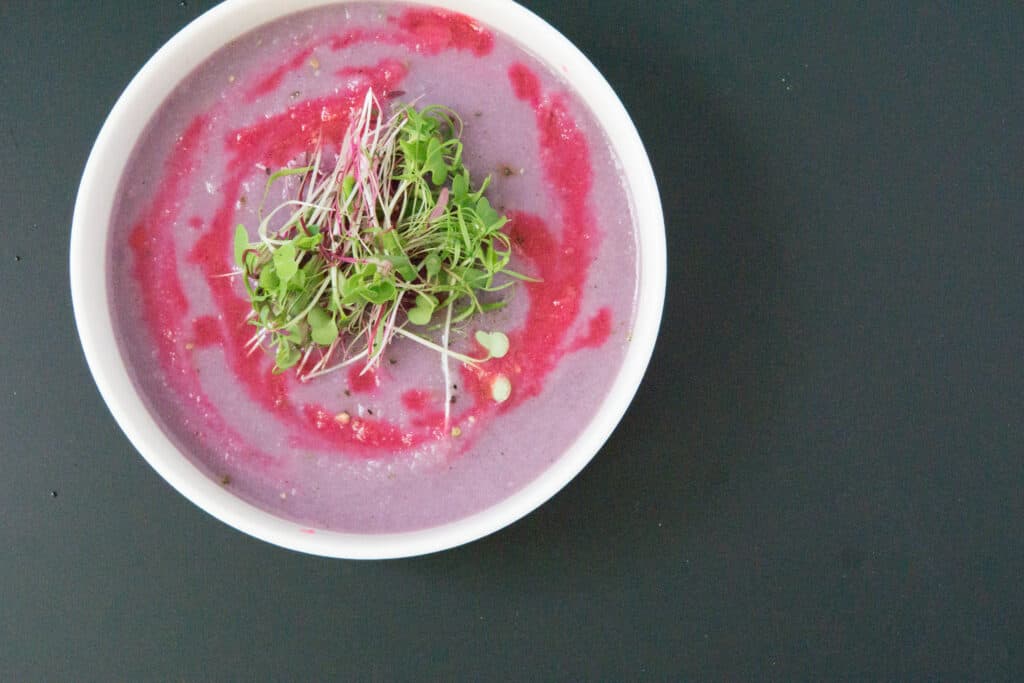 Purple Cauliflower Soup - Eat Your Way Clean 1 white bowl of bright purple soup with fuchsia drizzle and micro greens on black table