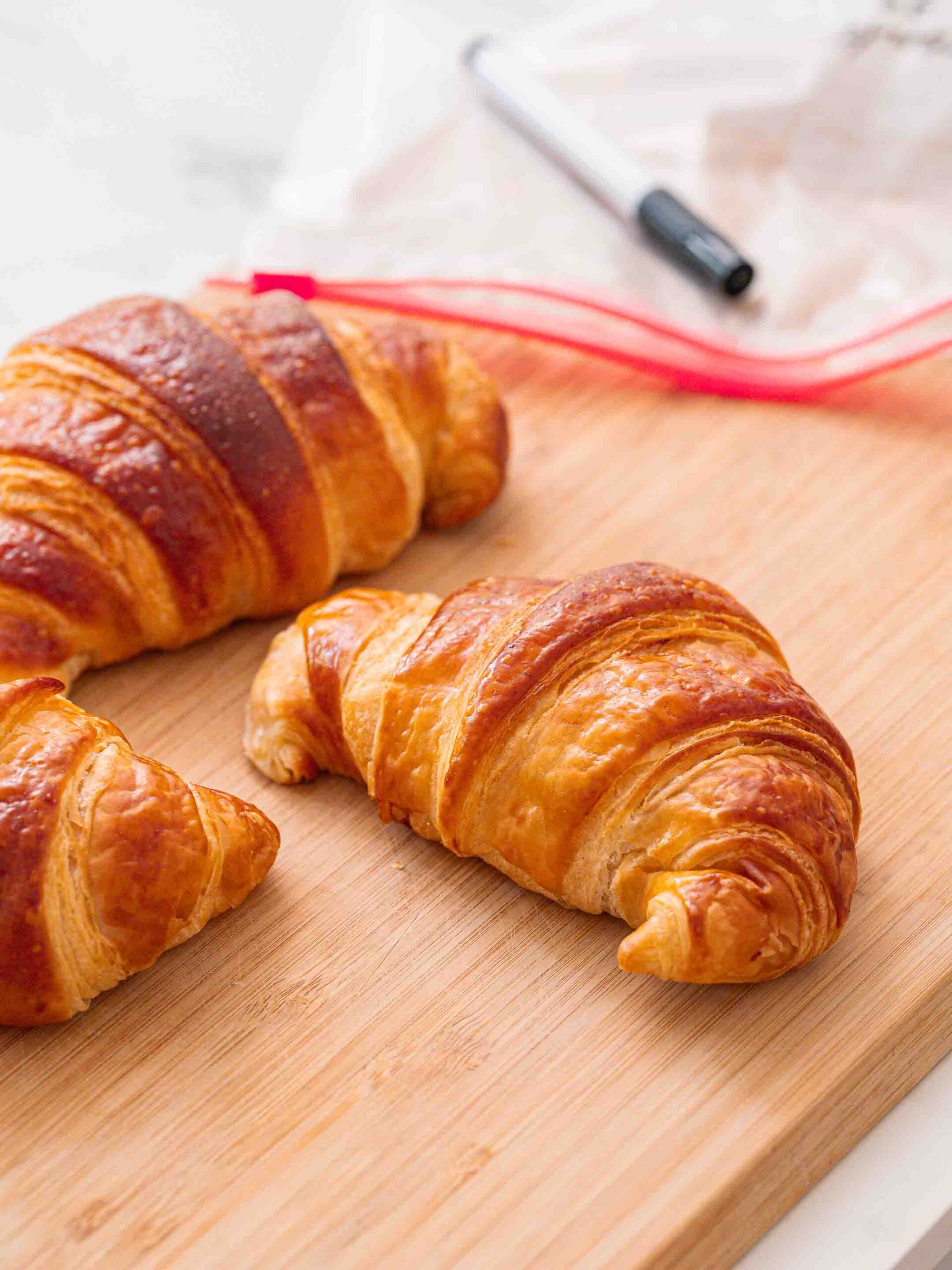 Three croissants on a wooden cutting board, with a black marker and a zipped plastic bag in the background.